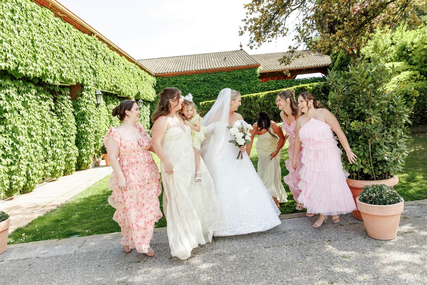 Bride in white gown walks with bridesmaids in floral dresses outside ivy-covered venue on sunny wedding day.