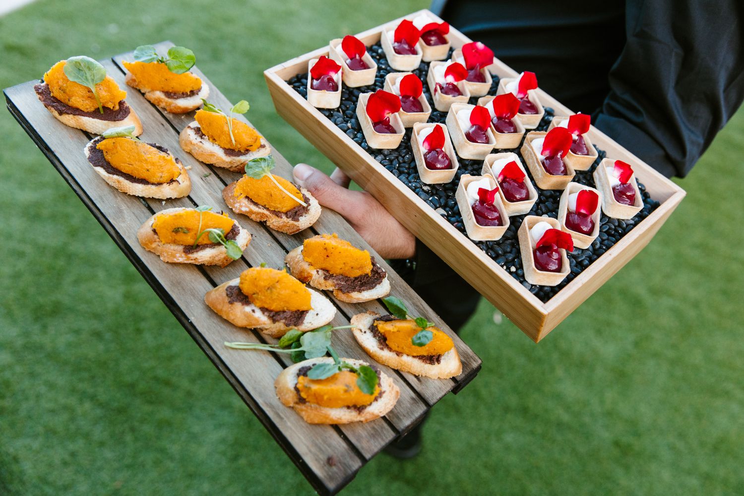 Server holding wooden trays of gourmet canapés, including orange-topped crostini with fresh herbs and bite-sized appetizers garnished with 
