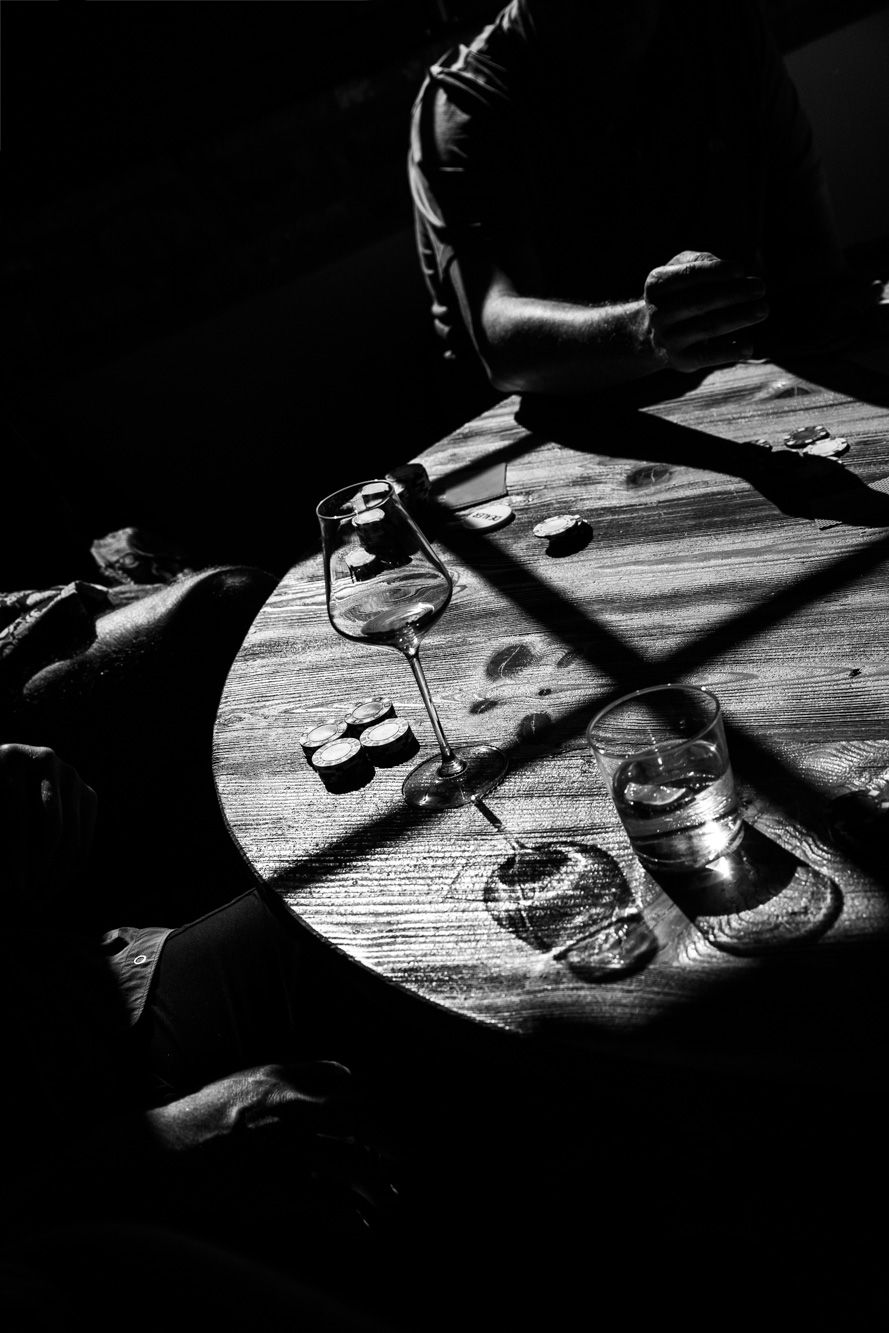 Black and white photo of a round wooden table with a wine glass, water glass and coins, lit by dramatic shadows as a person leans in.