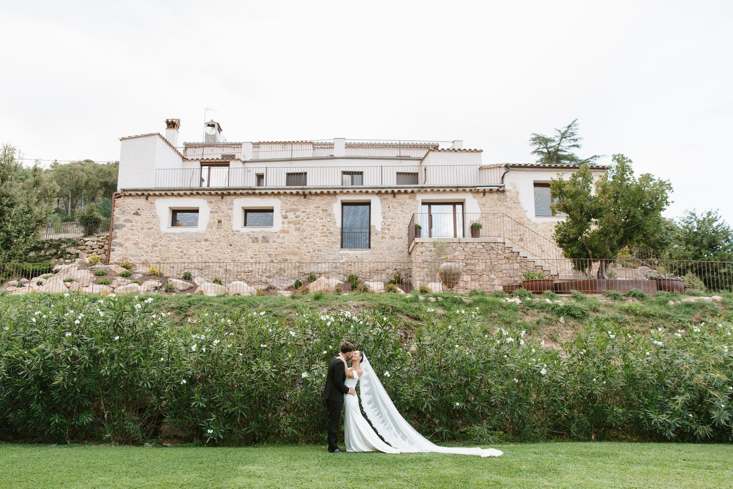 Bride and groom share a romantic kiss on lush green lawn before a rustic stone villa wedding venue.