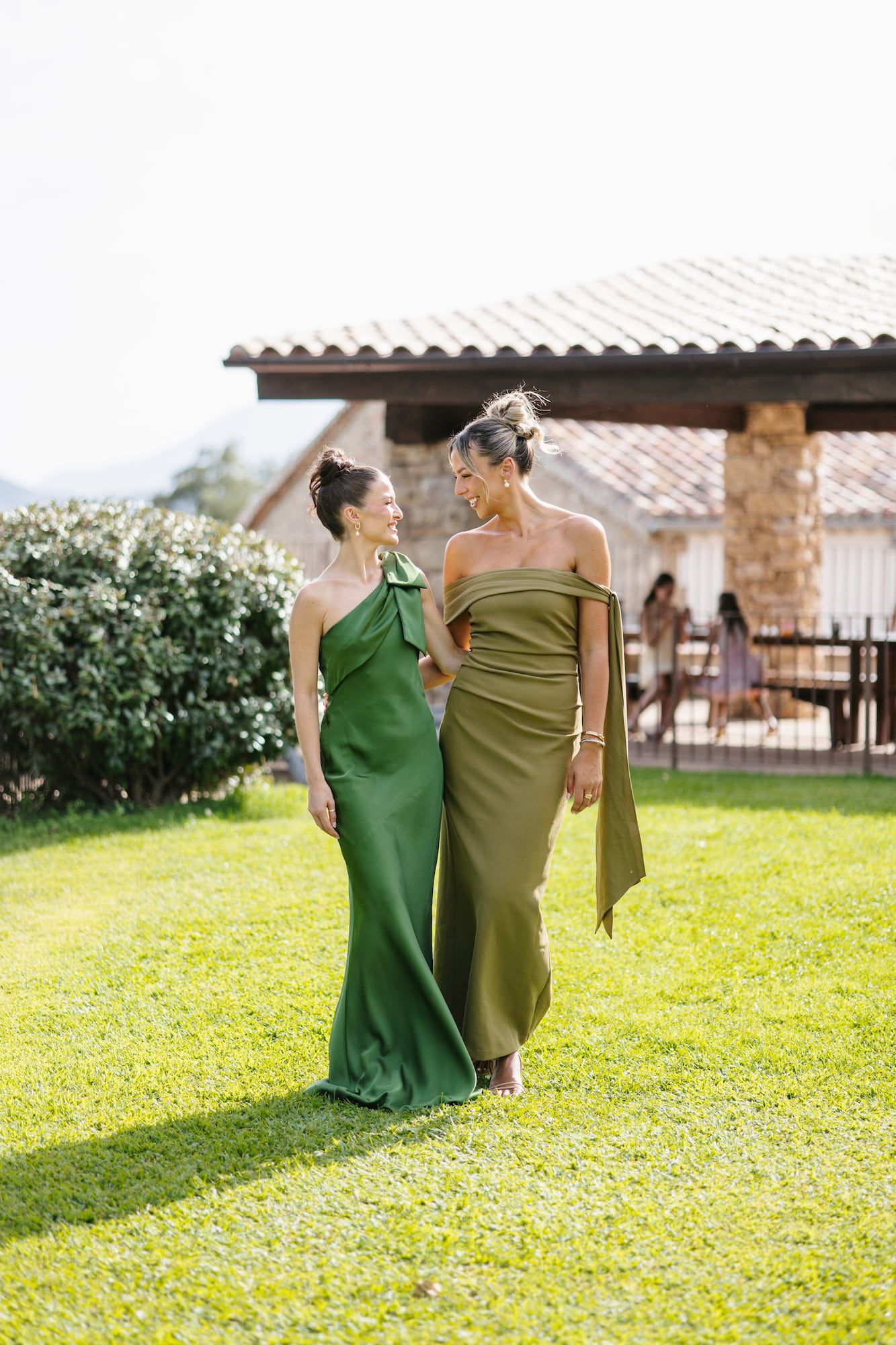 Two women in elegant green gowns walk together on a lawn at an outdoor venue, smiling at each other.