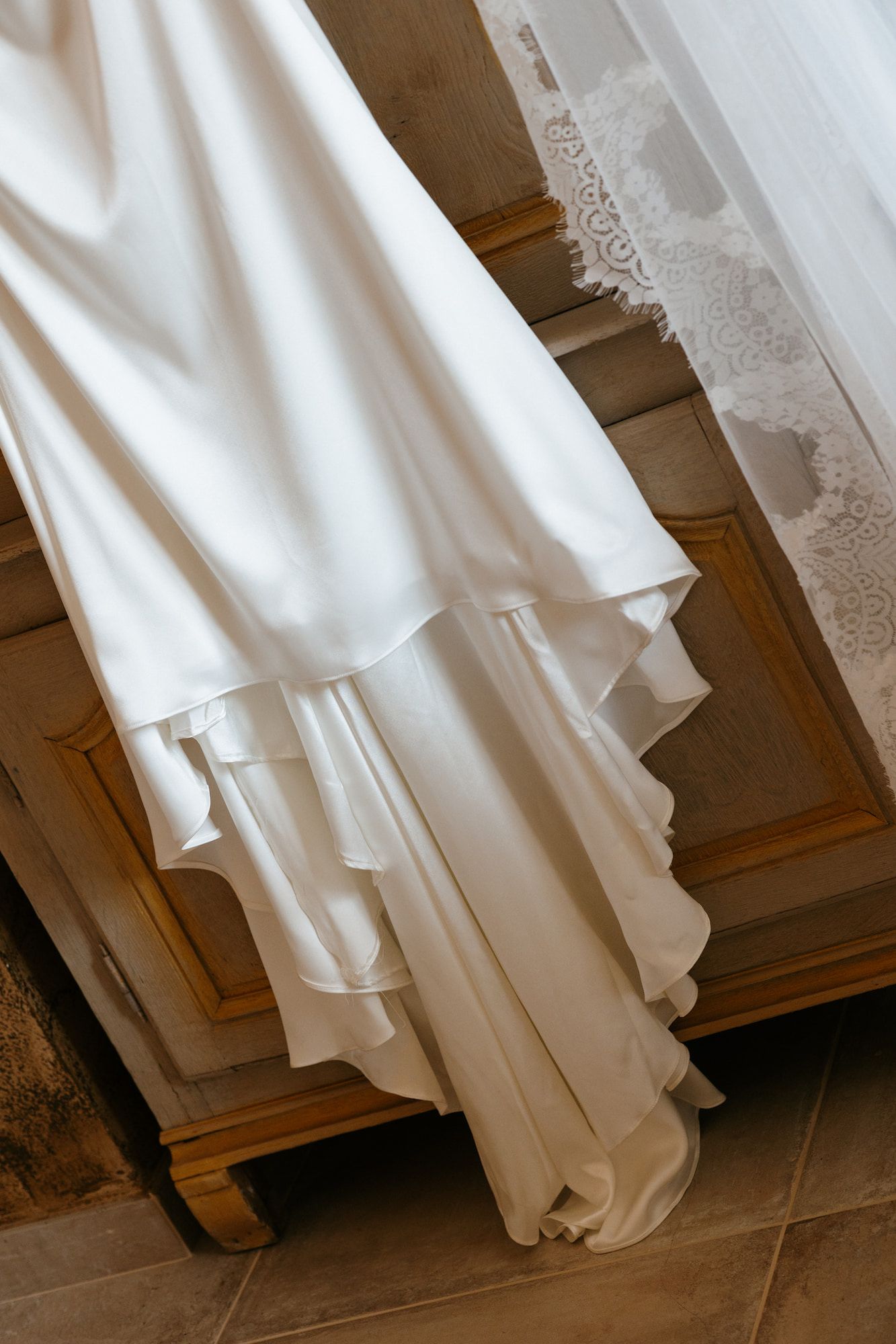 White satin wedding dress with ruffled hem draped over a wooden cabinet with lace veil detail.
