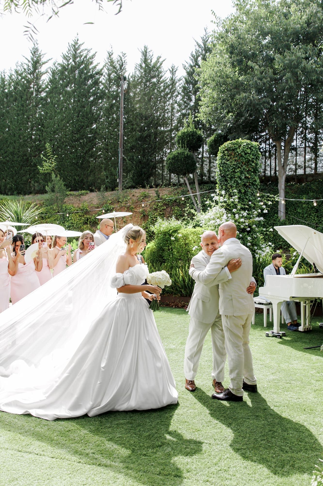 Bride in a flowing white gown stands with two men in a lush garden wedding ceremony outdoors.