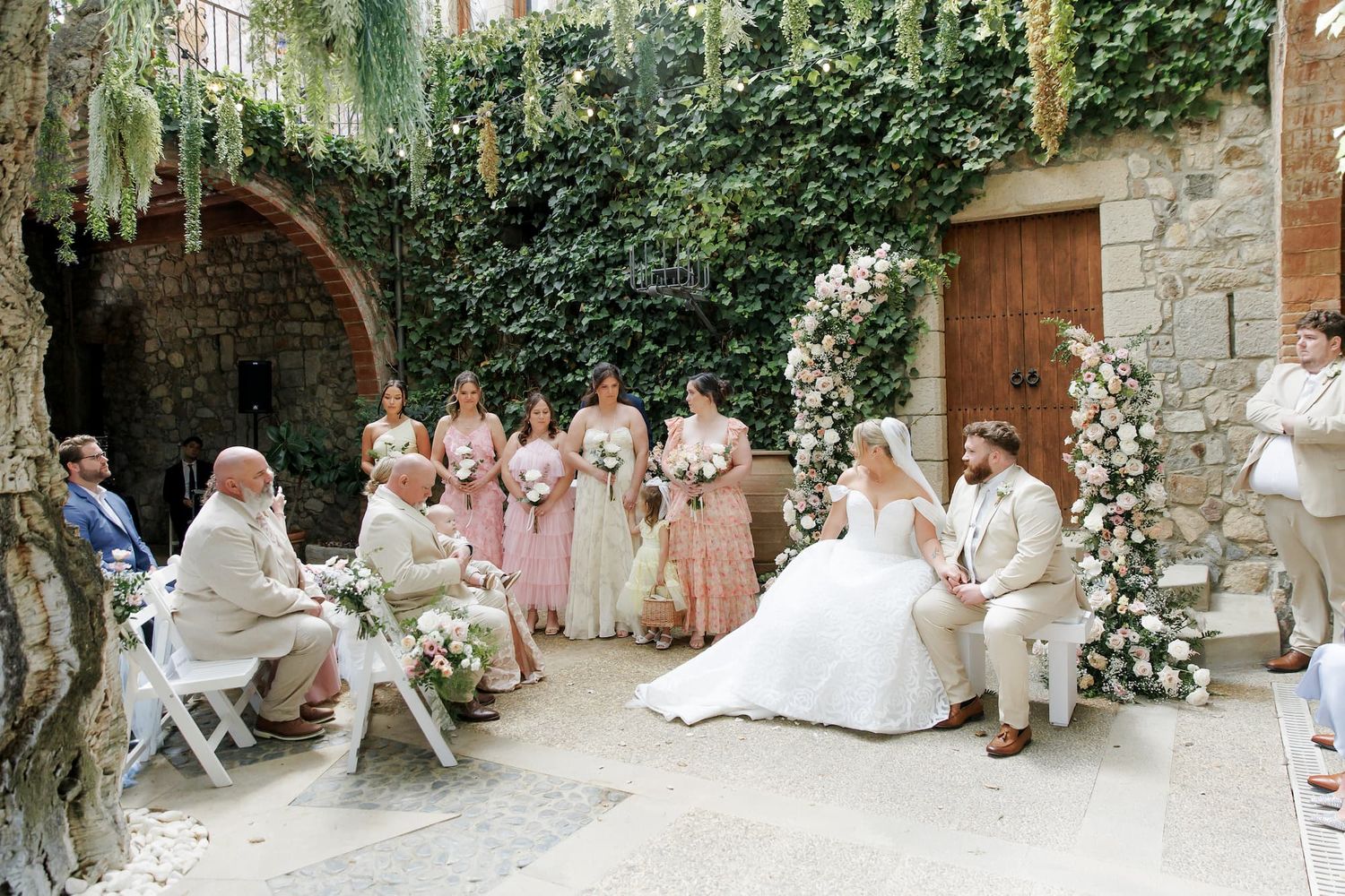Outdoor wedding ceremony in an ivy-covered courtyard with bride, groom, bridesmaids in pink, and guests seated on white chairs.