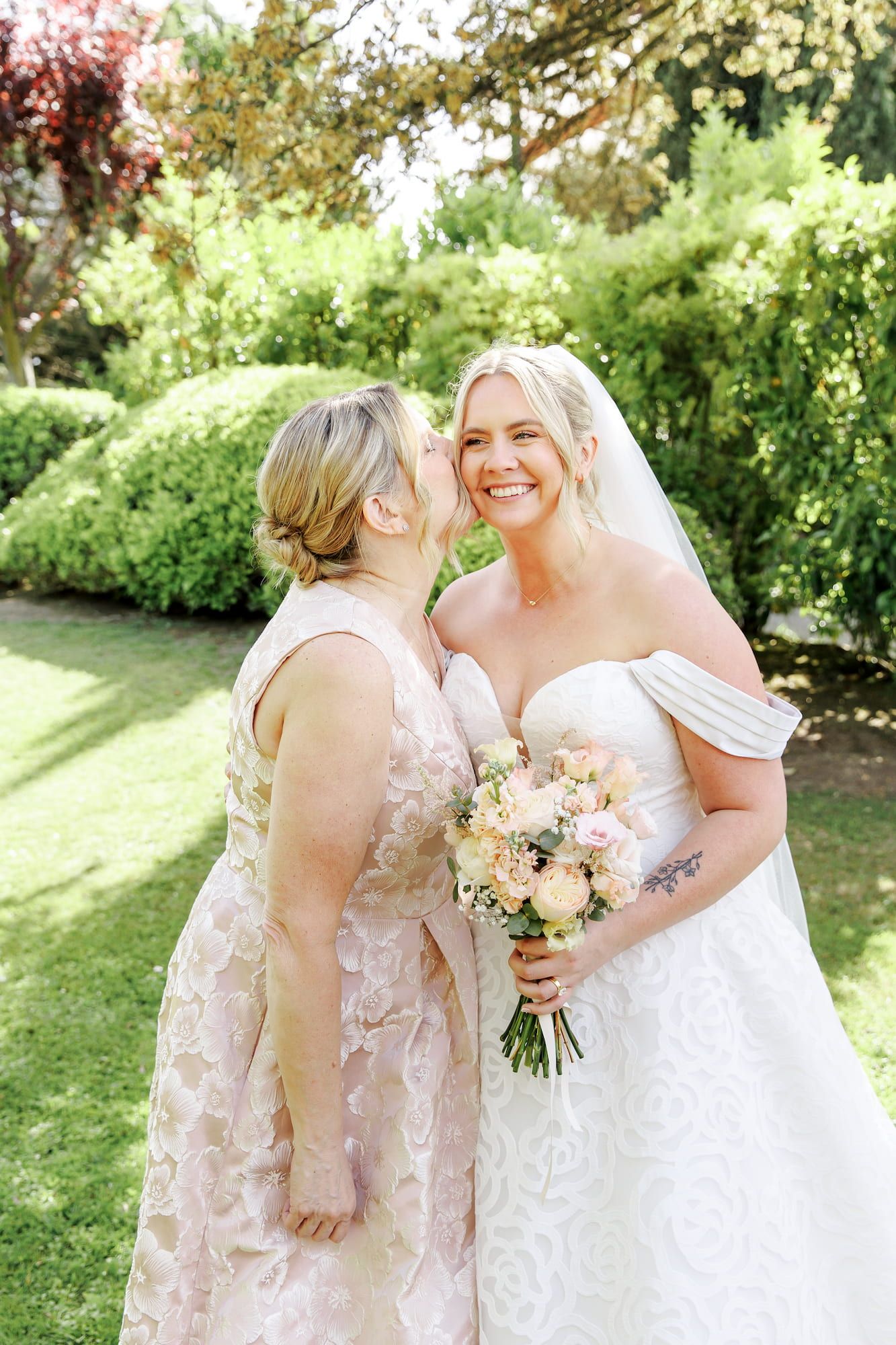 Two brides share a tender moment outdoors, one in a white gown with veil holding a pastel bouquet, the other in a floral dress.