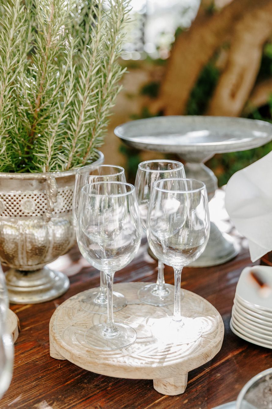 Empty wine glasses on a wooden board beside silver planters with rosemary at an elegant outdoor table setting.