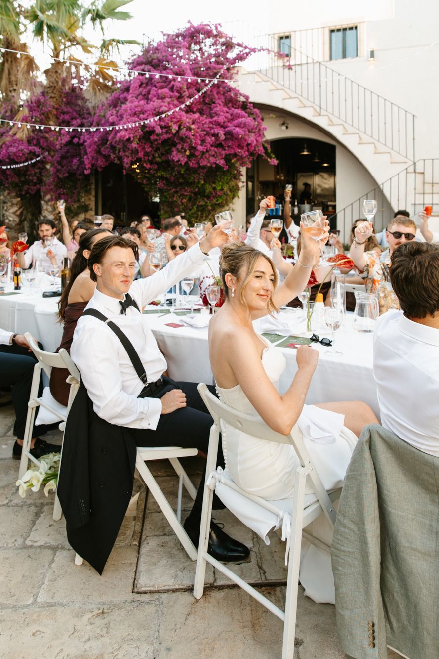 Bride and groom toast with guests at outdoor wedding reception under blooming pink bougainvillea in Mediterranean venue.