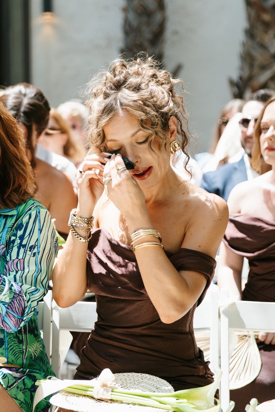 Emotional wedding guest in brown dress wiping tears during outdoor ceremony.