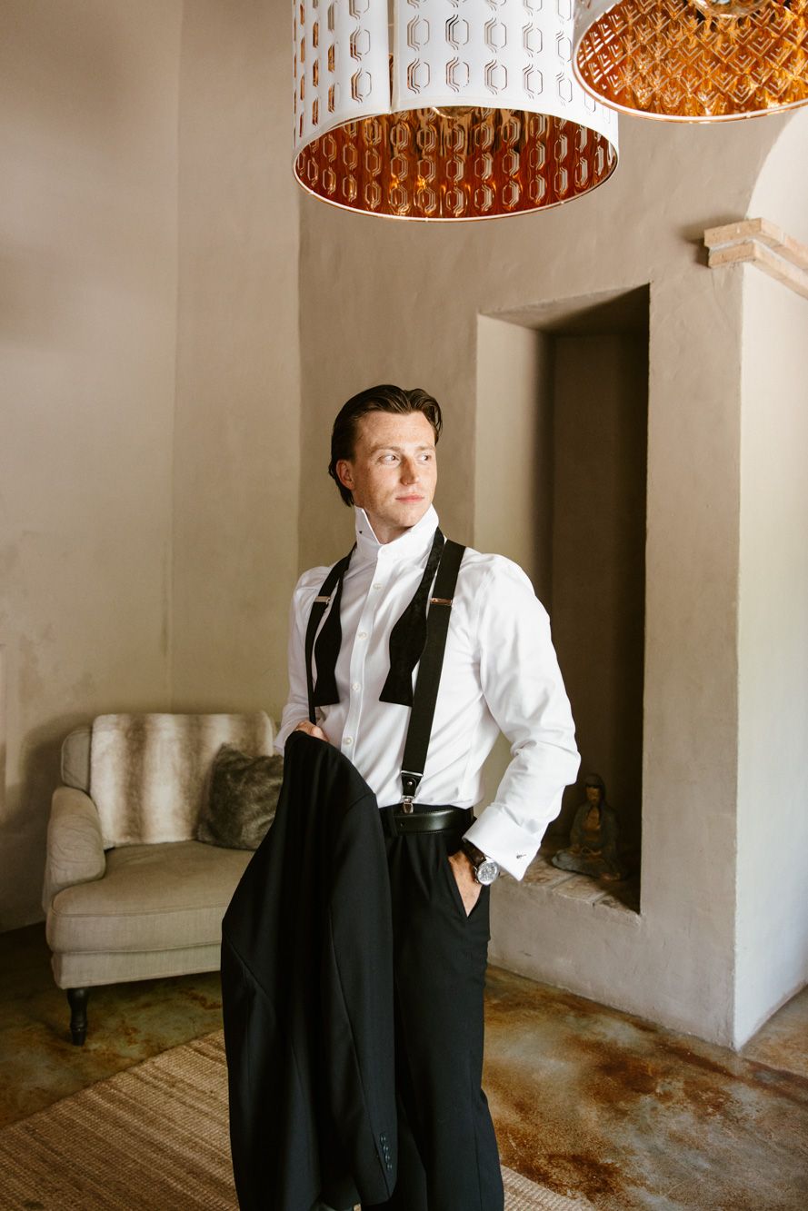 Elegant man in white shirt with suspenders holding black trousers in a stylish room with copper pendant lights.