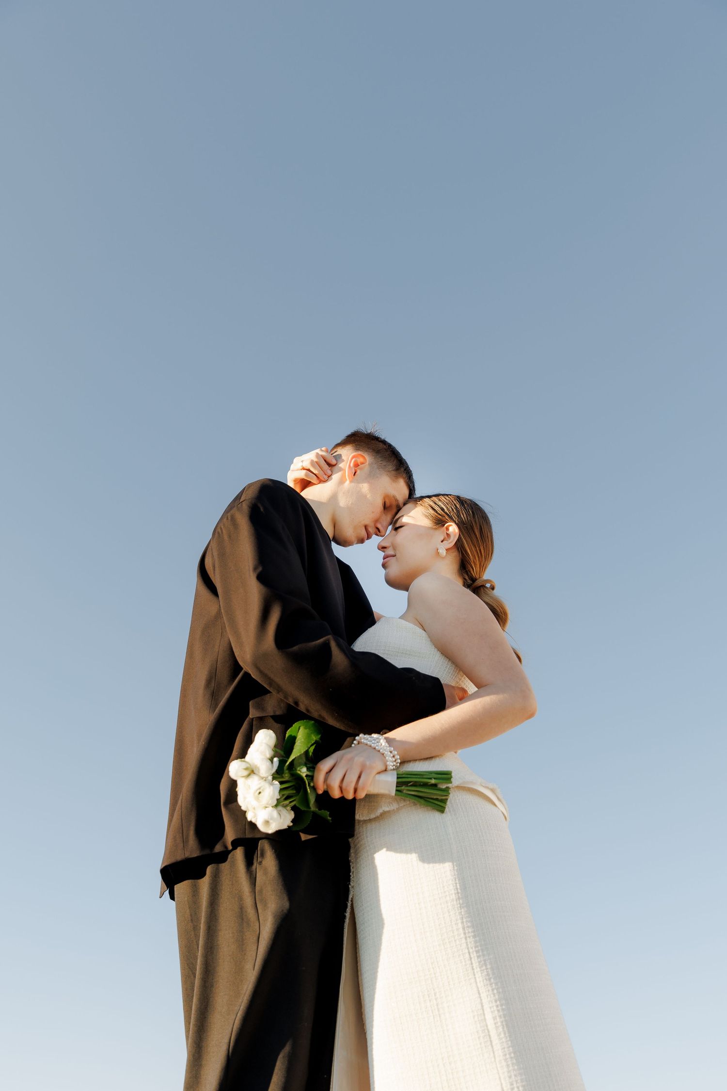 A bride and groom share a romantic forehead touch moment outdoors, bride holding white bouquet against clear blue sky.