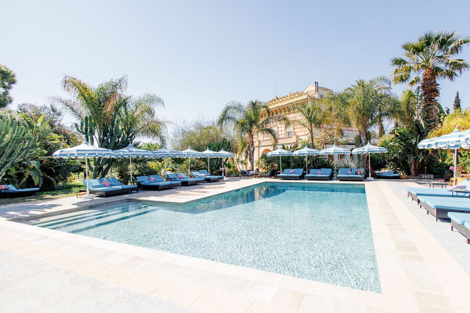 Luxury outdoor pool with white umbrellas, lounge chairs, palm trees, and an elegant villa in the background.
