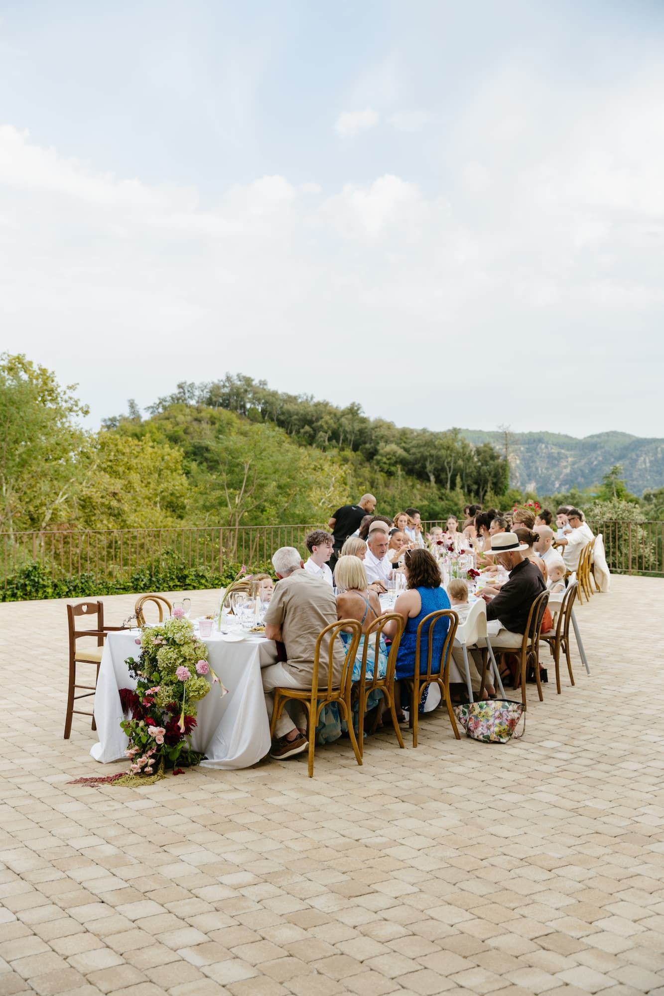 Guests dining at a long outdoor table on a stone terrace with lush green mountain views at a wedding reception.