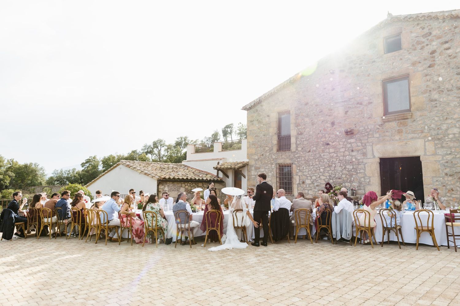 Outdoor wedding reception at a rustic stone venue with guests seated at long tables under open sky.