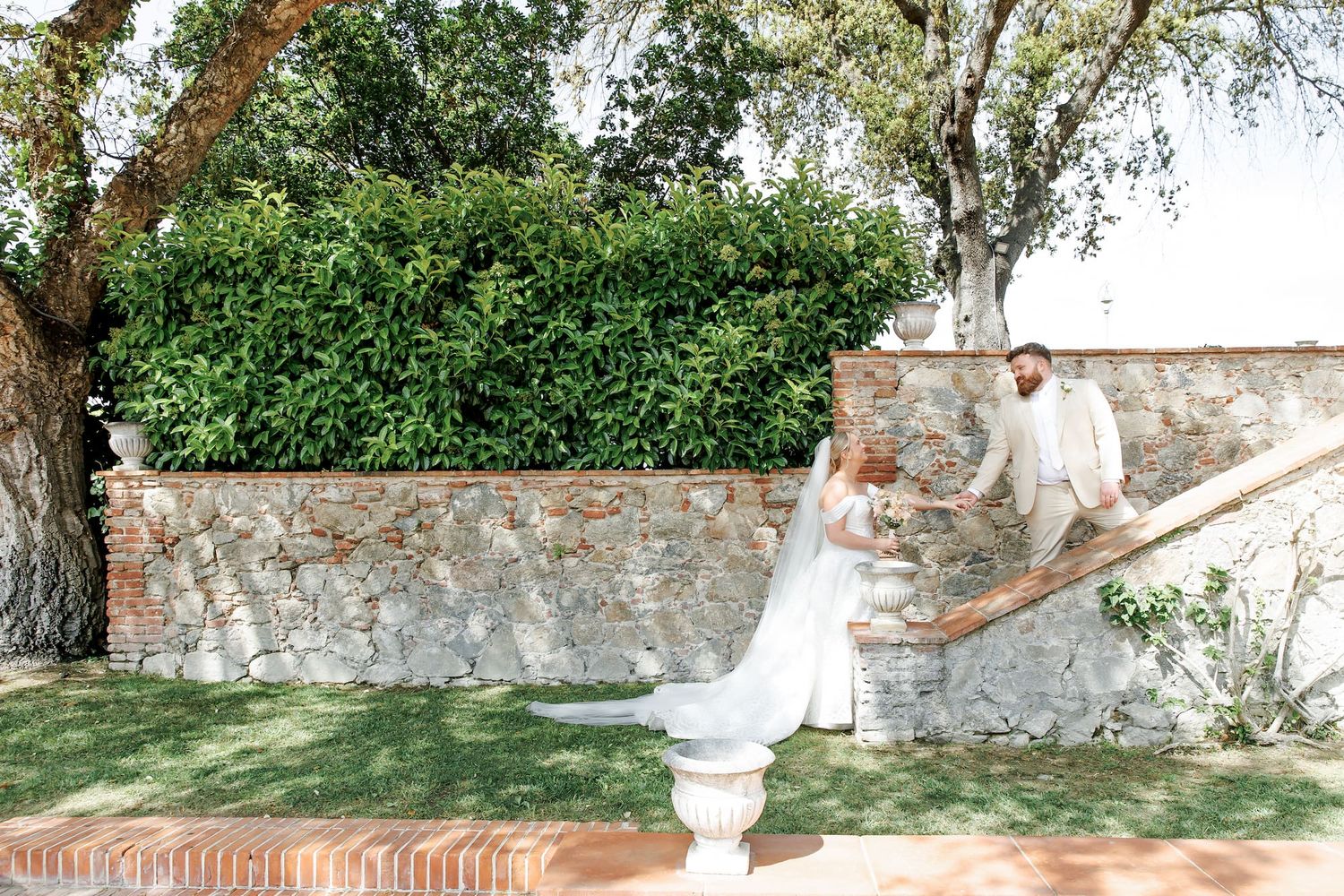 Bride and groom walk hand in hand in a sunlit garden with stone wall and lush greenery.