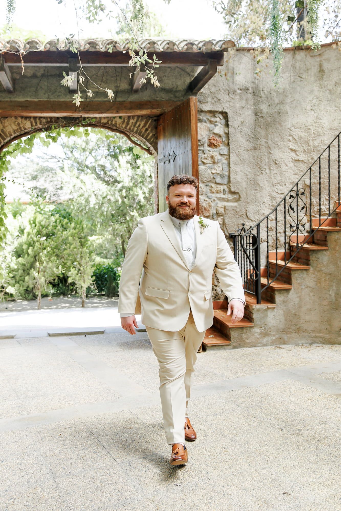Groom in beige suit walks confidently near stone archway and staircase at outdoor wedding venue.