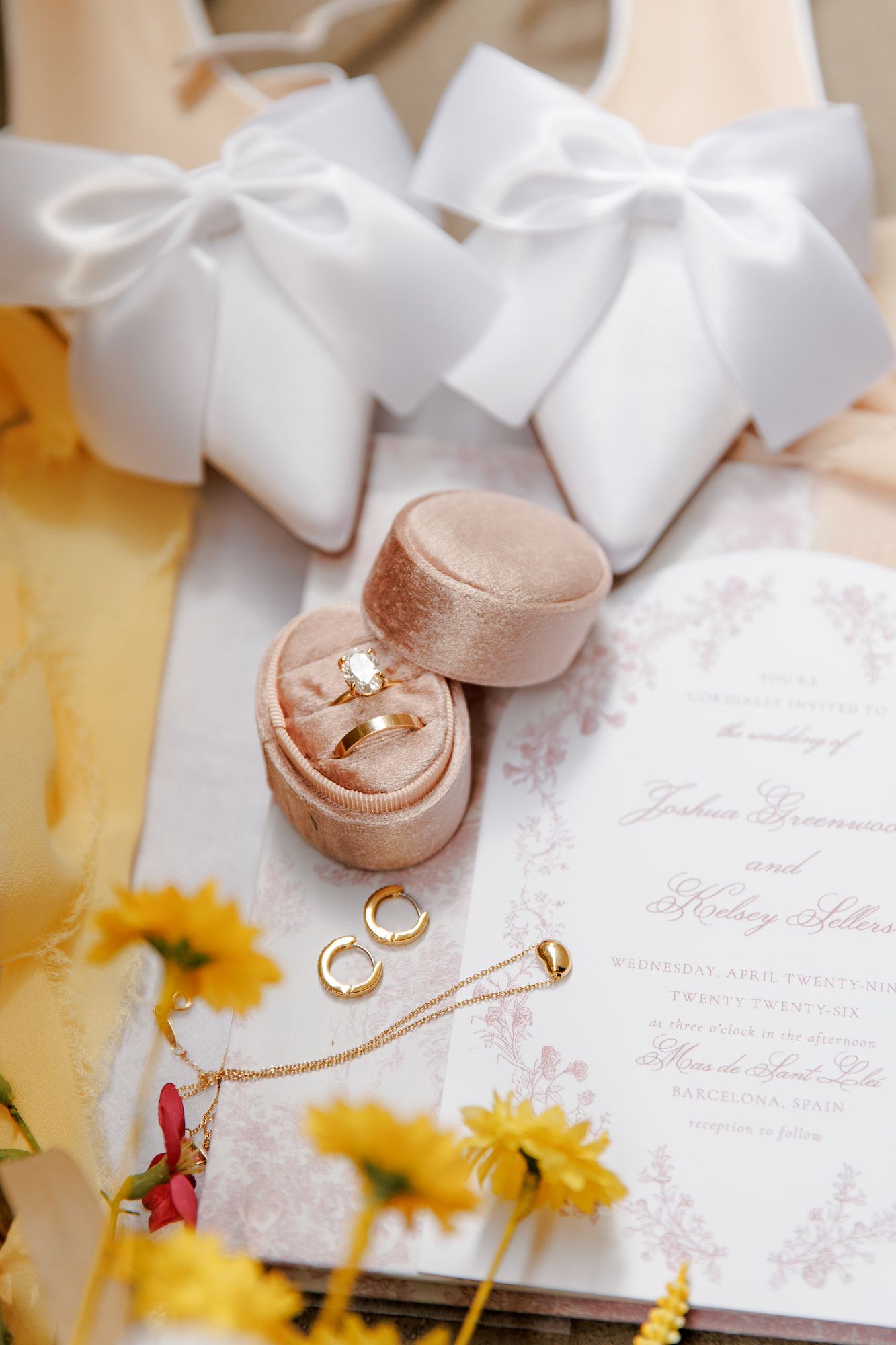Elegant wedding flat lay with gold rings in pink velvet box, floral invitation, yellow flowers, and white bow accessories.