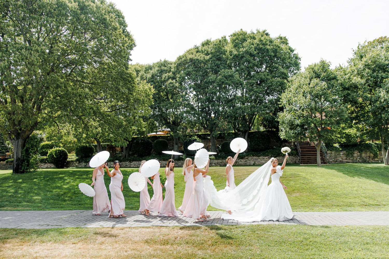 Bride and bridesmaids in pink dresses holding white parasols on a sunny green lawn at an outdoor wedding.