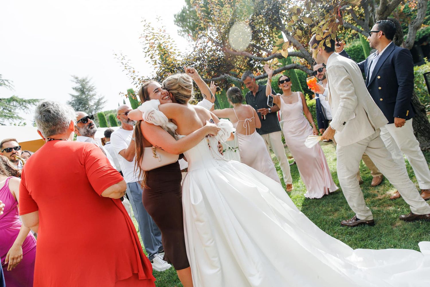 Bride in white gown hugging guests outdoors at a wedding celebration with bridesmaids and attendees.