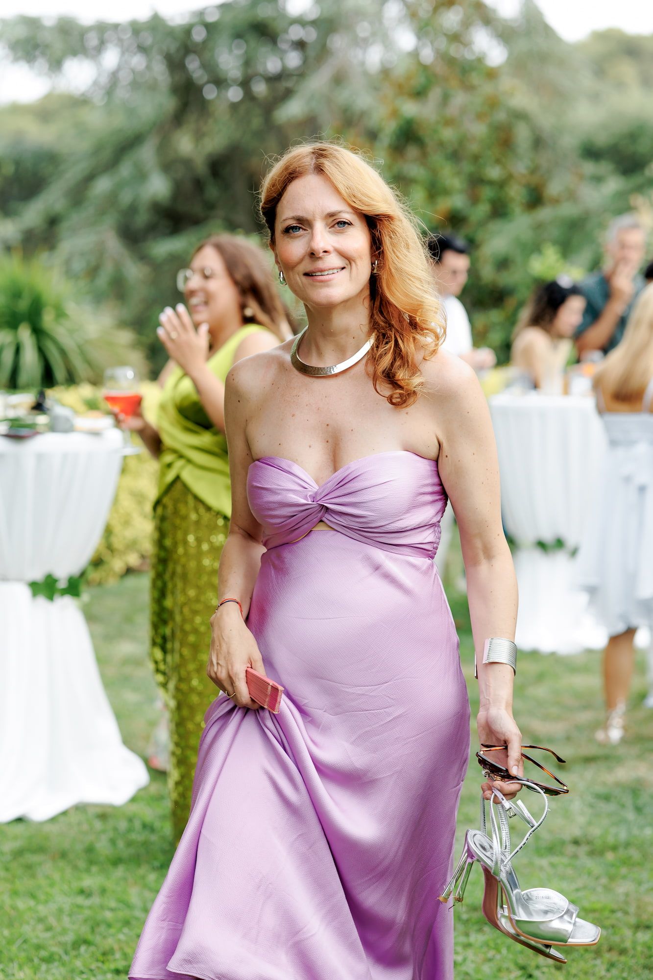 Woman in lavender satin gown holding silver heels at outdoor garden party event.