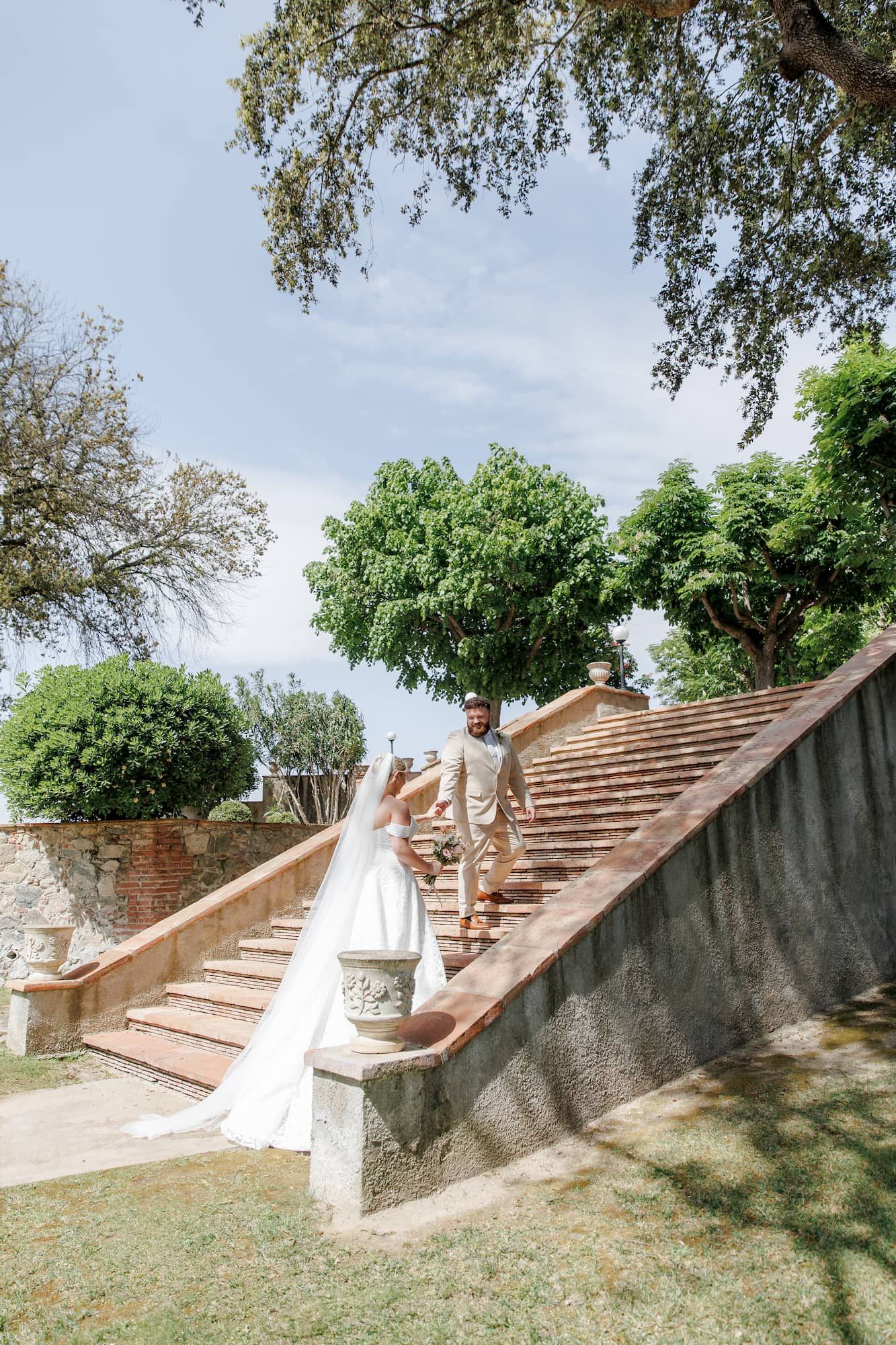 Bride and groom walking up stone stairs surrounded by lush green trees at an outdoor wedding venue in Barcelona