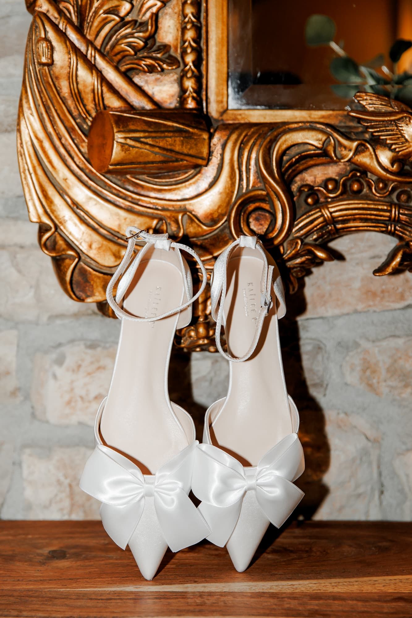 White bridal heels with satin bow details hanging on ornate gold mirror frame.