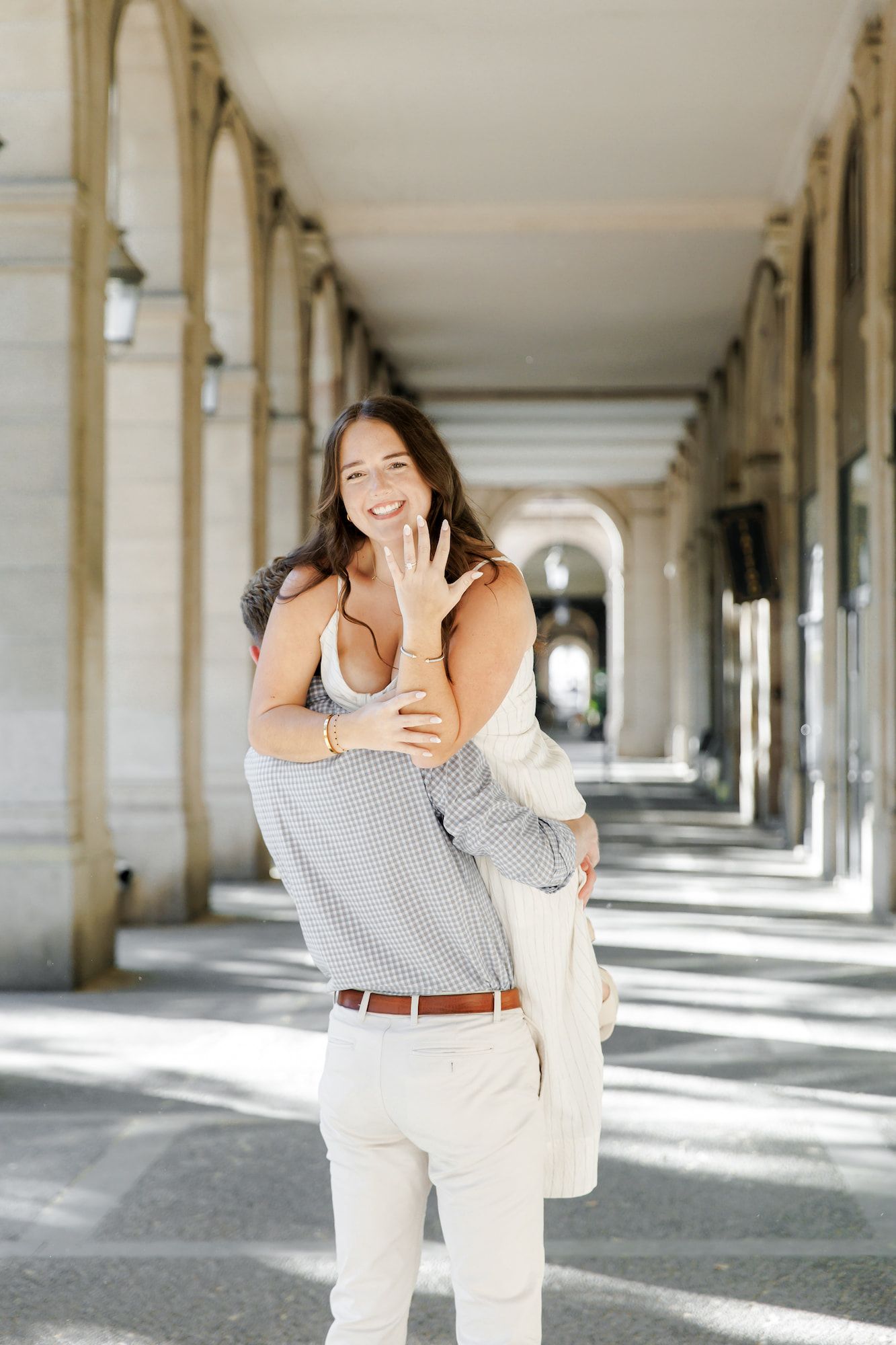 Happy woman shows off engagement ring while being carried by partner in sunlit European arcade colonnade.