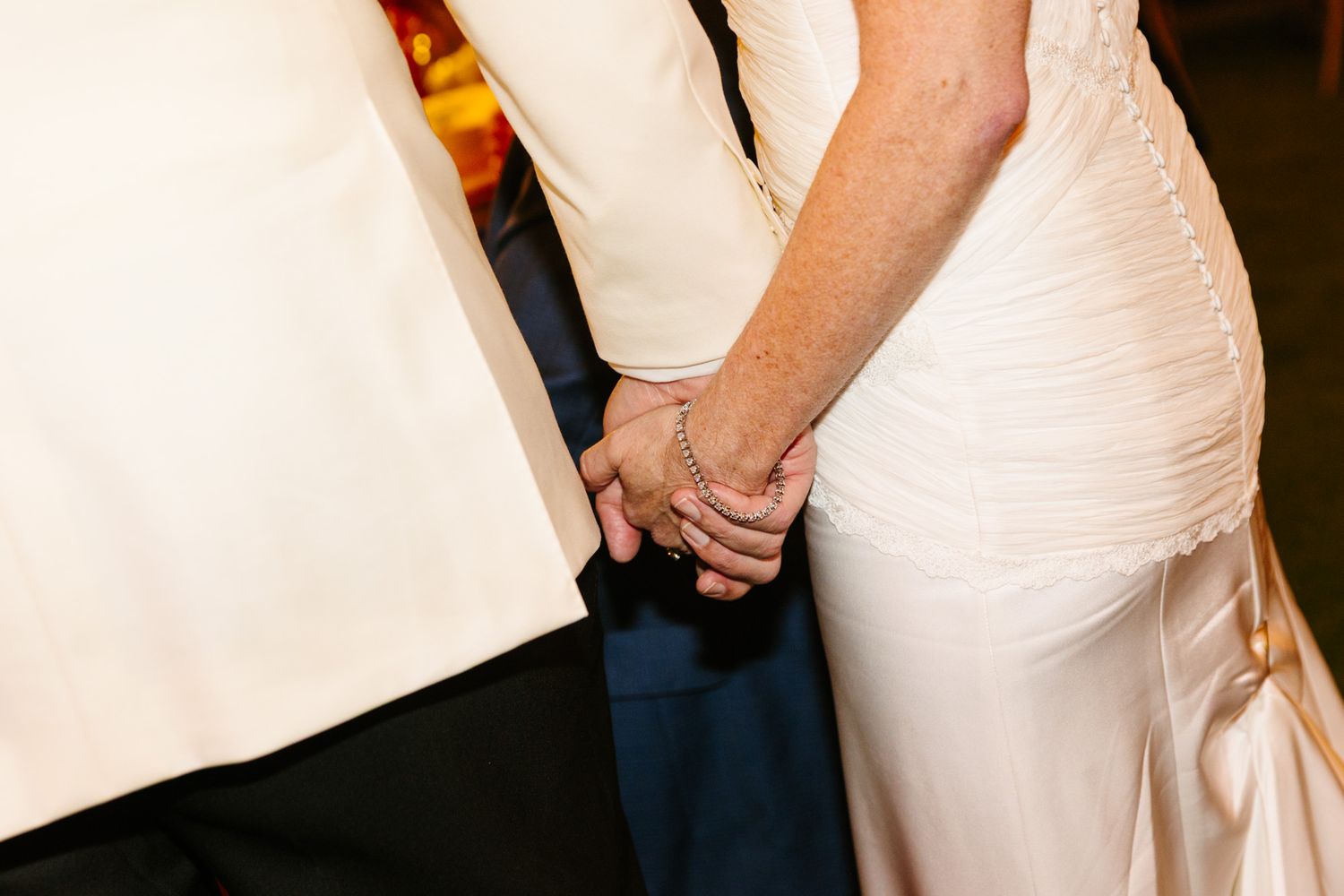 Bride in a white lace wedding gown holds hands with a groom in a cream tuxedo jacket during a wedding celebration.