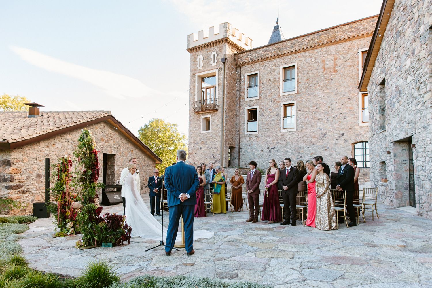 Wedding ceremony on a stone patio at a medieval castle venue, with the officiant, bride, groom and guests in colorful dresses watching.