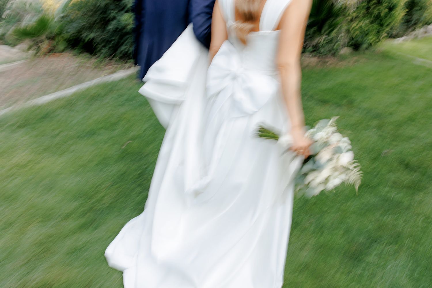 A bride in a flowing white wedding dress twirls gracefully on a grassy lawn in a garden setting.