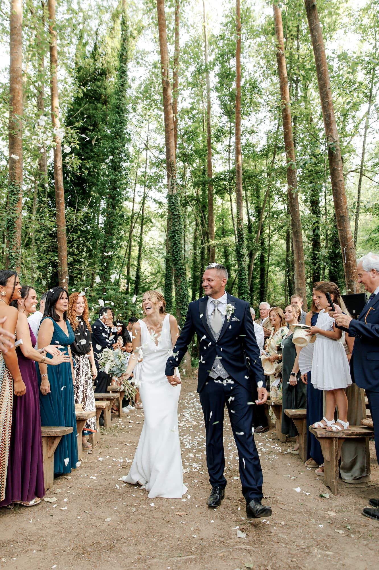 Bride and groom walk down forest aisle as guests throw white confetti at an outdoor woodland wedding ceremony.