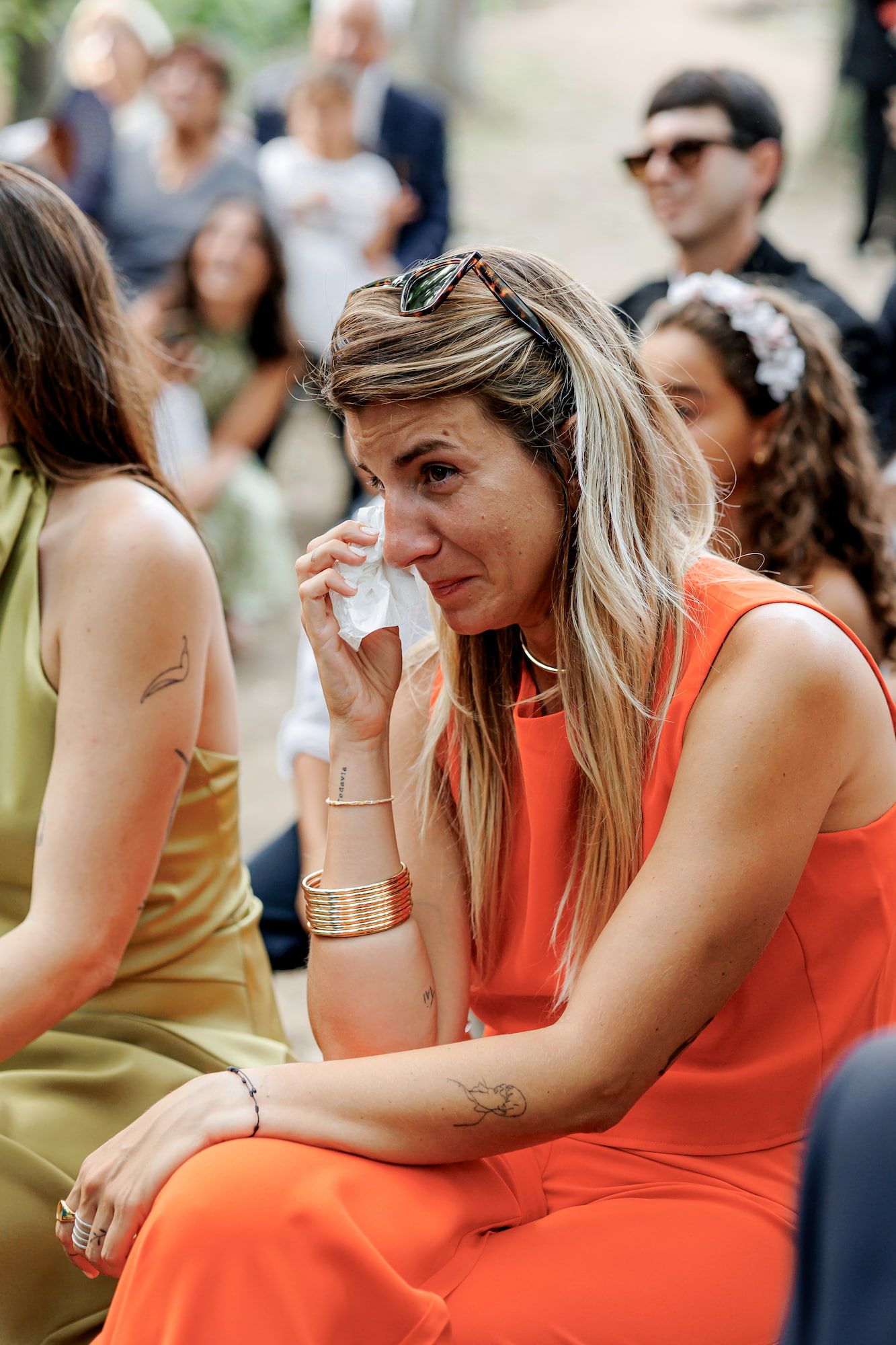 Woman in orange dress crying and wiping tears with tissue at an outdoor wedding ceremony.