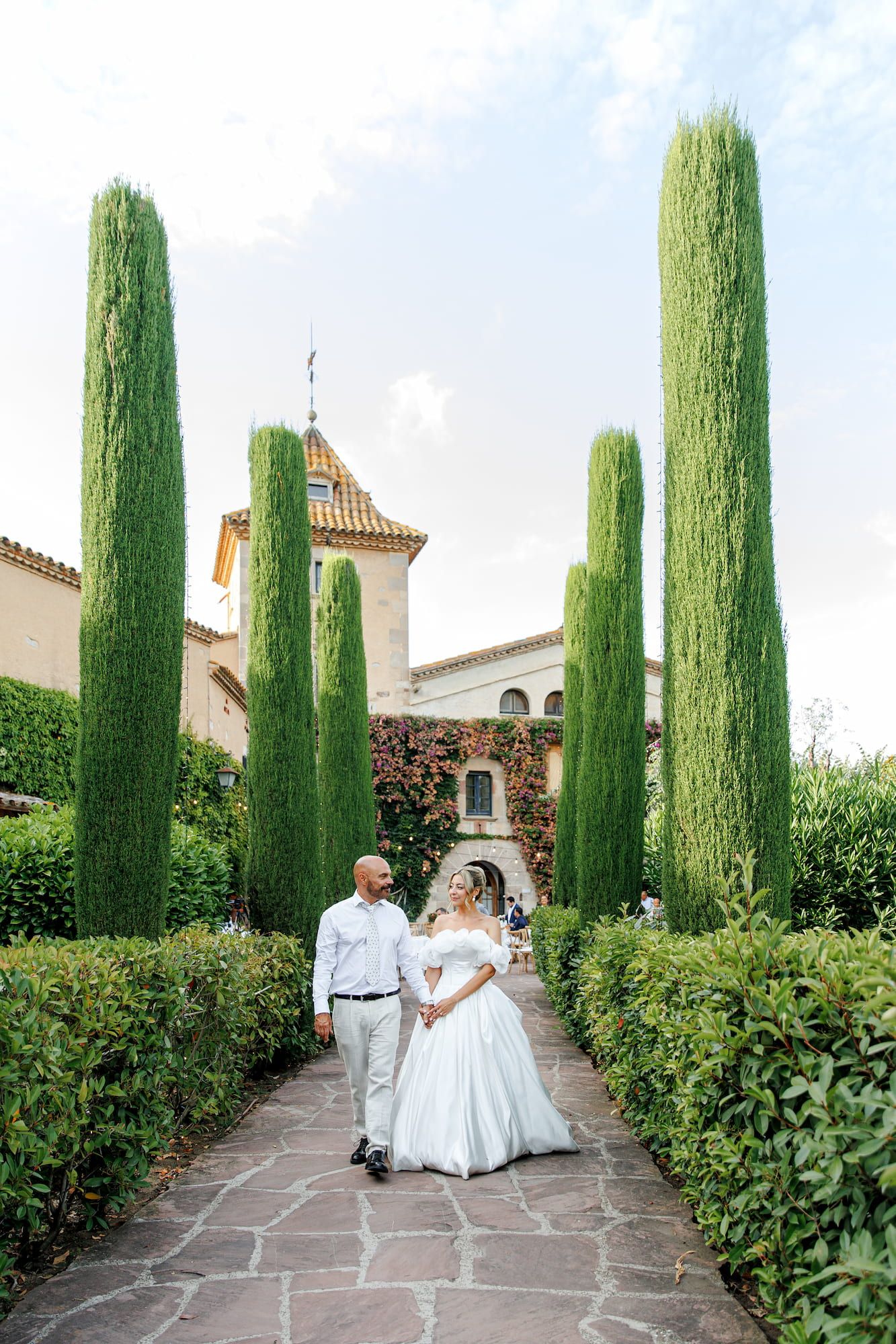 Bride and groom walk hand-in-hand through a lush garden path lined with tall cypress trees at a Mediterranean villa.