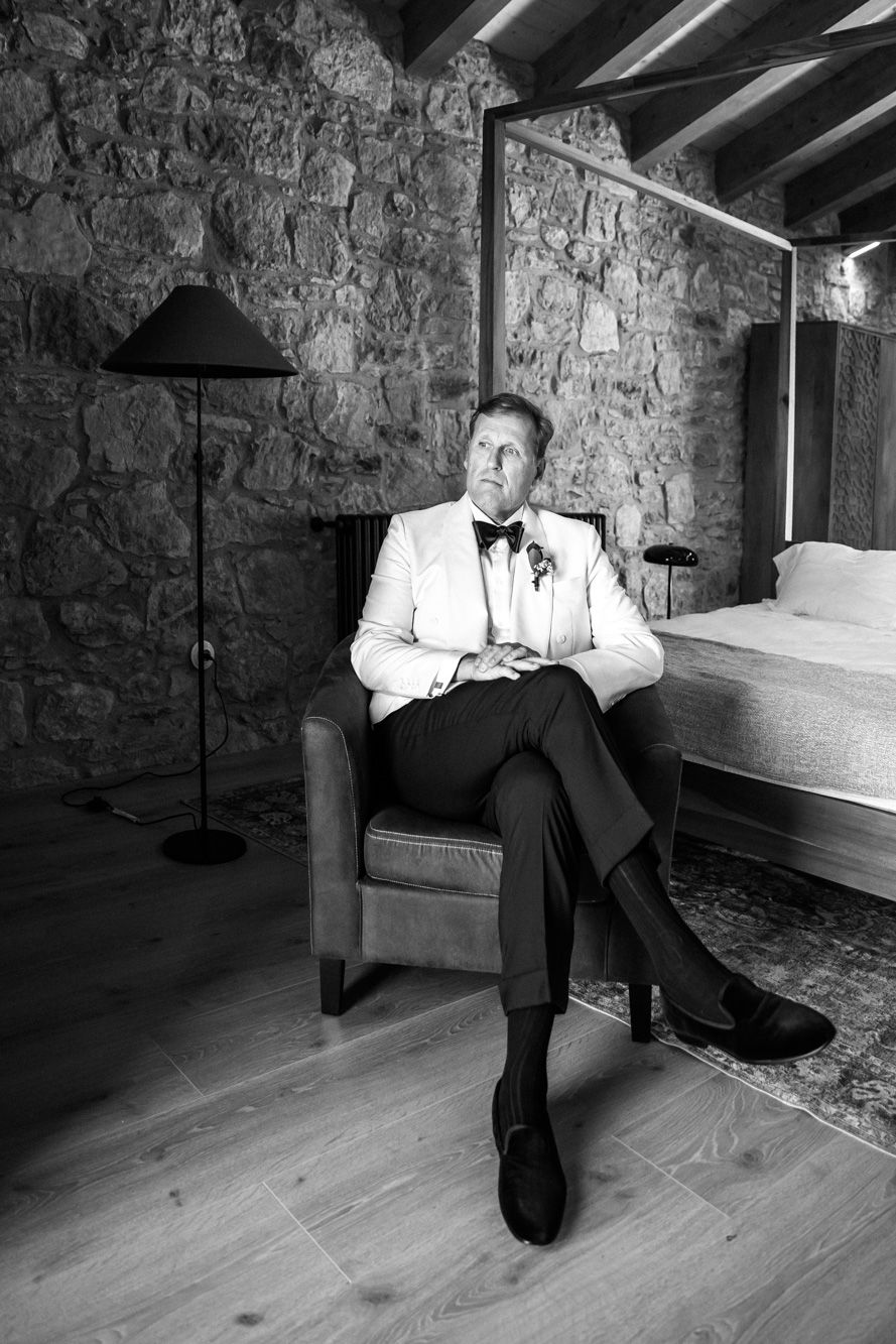 Groom in a white dinner jacket and bow tie sits in a leather armchair in a rustic stone-walled bedroom with a canopy bed.