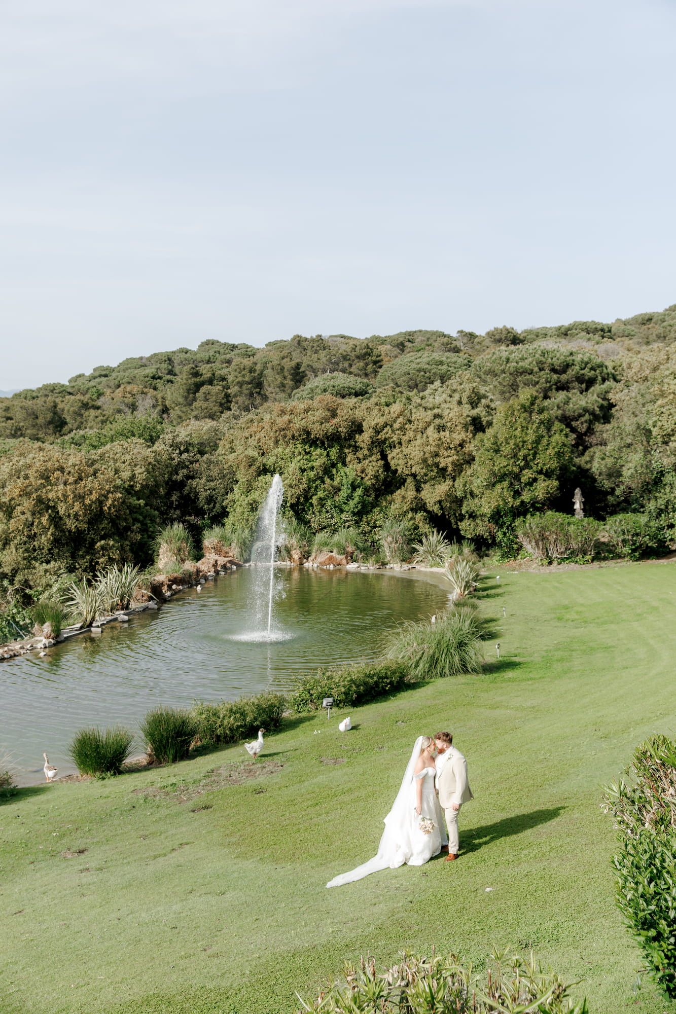 Bride and groom share a romantic moment on a lush green lawn beside a serene pond at Mas de Sant Lleí.