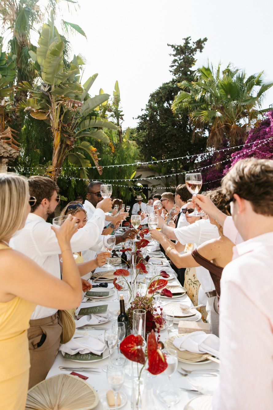 Guests raising wine glasses in a toast at an outdoor garden wedding reception with white decor and tropical greenery.