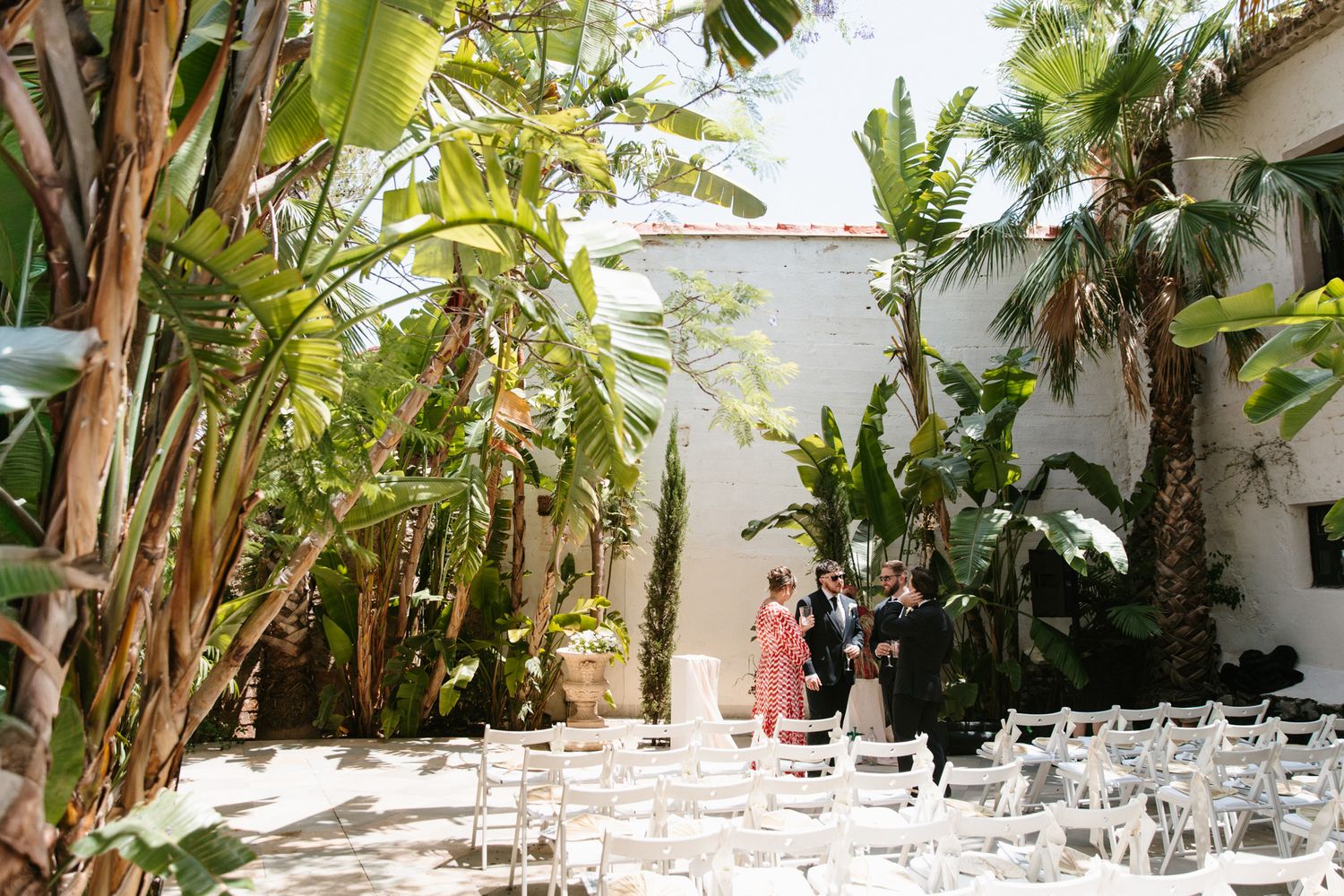 Outdoor wedding ceremony setup with white chairs and lush tropical palm and banana trees in a sunlit courtyard.