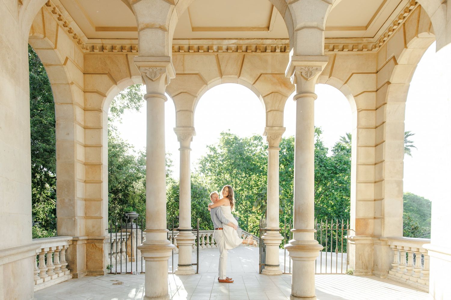 Couple embracing under elegant white stone arches and columns at a classical outdoor pavilion surrounded by lush greenery.