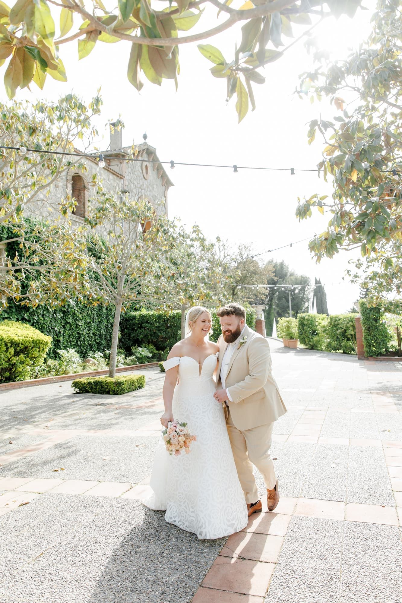 Bride in white lace gown and groom in tan suit walk together outdoors at a sunlit garden wedding venue.