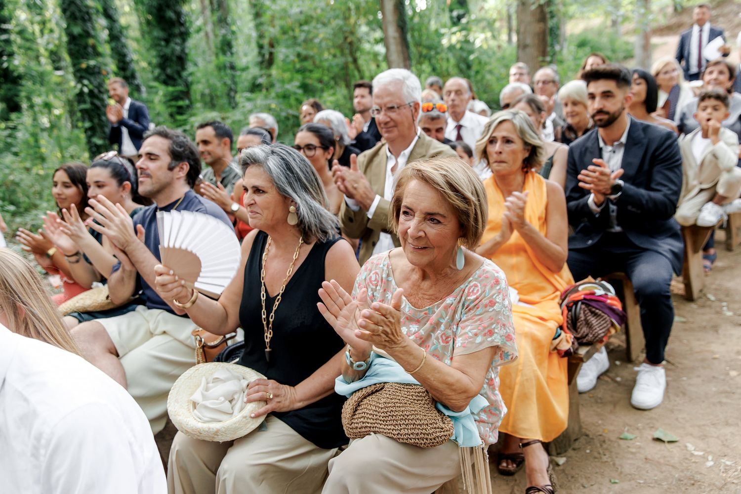 Guests clapping and smiling at an outdoor wedding ceremony seated among lush green trees.