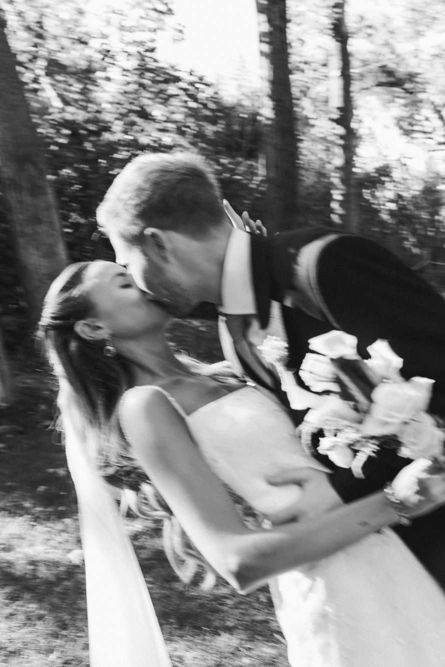 Black and white photo of a bride and groom sharing a romantic kiss outdoors, bride holding a bouquet.