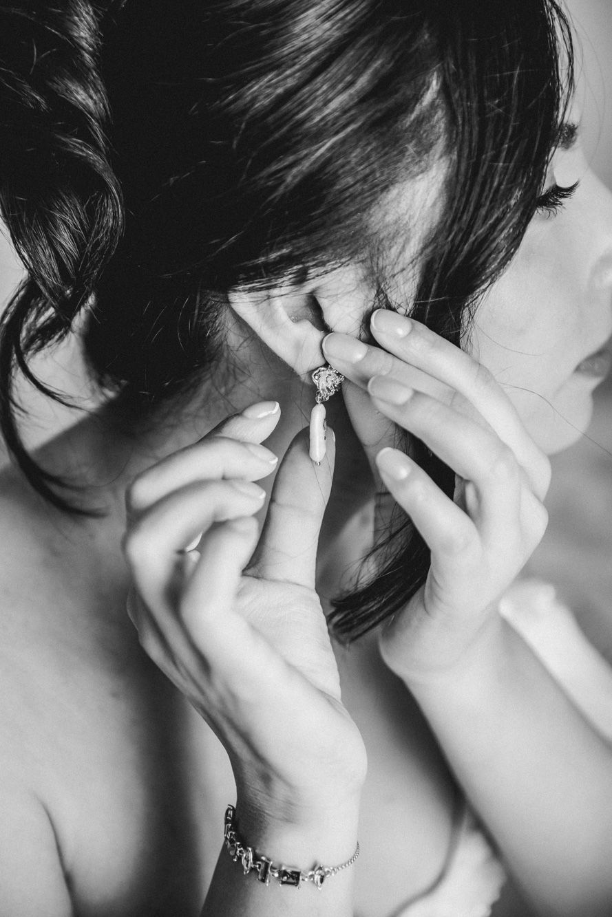 Bride putting on elegant drop earring in black and white photo, wearing bracelet, hair up for wedding day.