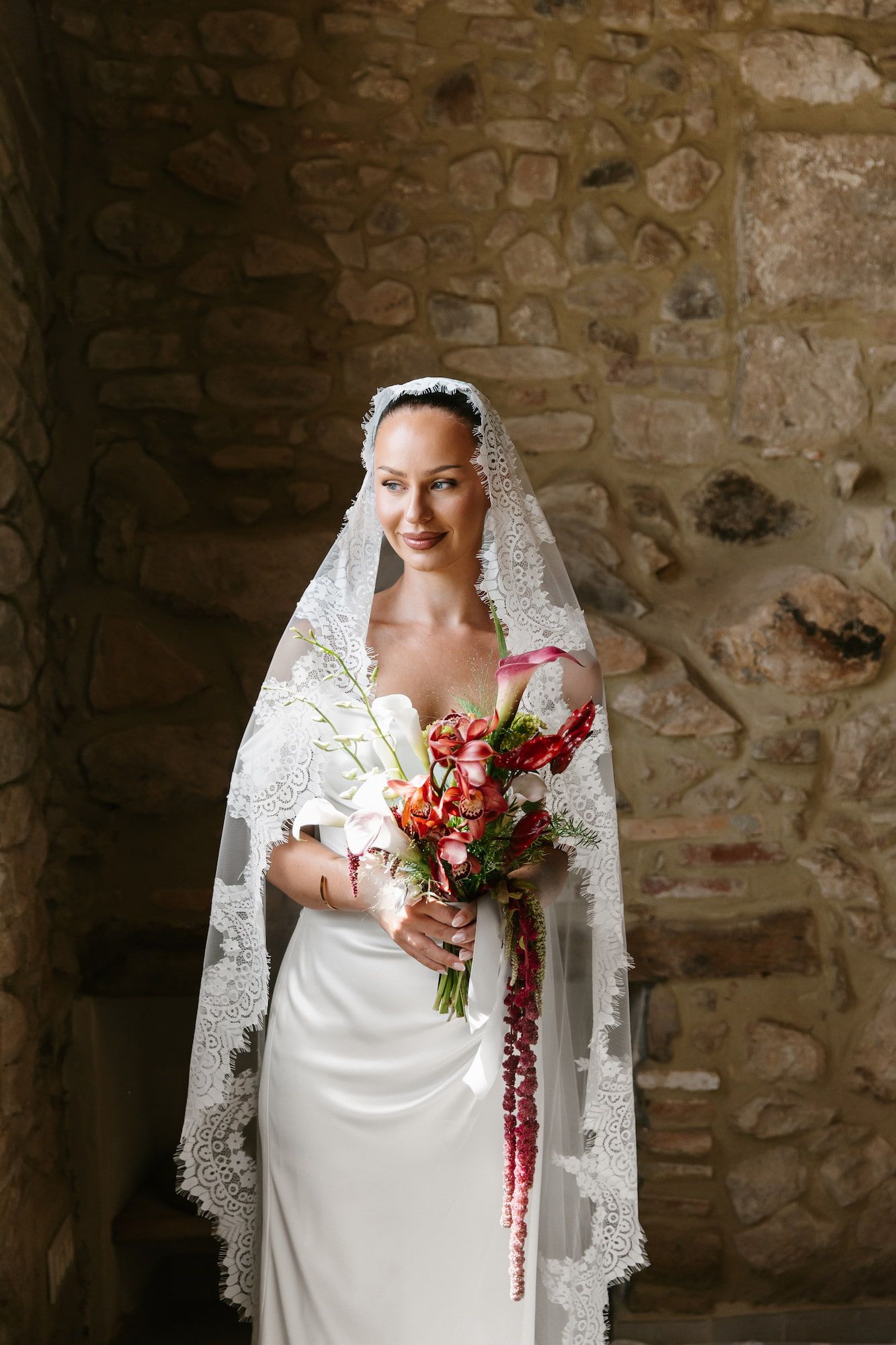 Elegant bride in white lace veil holding colorful bouquet, standing against rustic stone wall background.