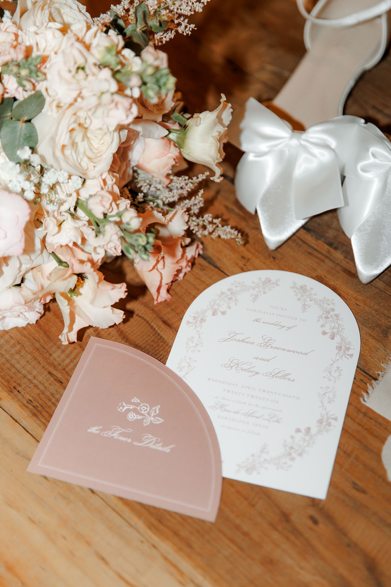 Elegant wedding flat lay with blush floral bouquet, arched invitation, pink program, and white bow heels on wood.