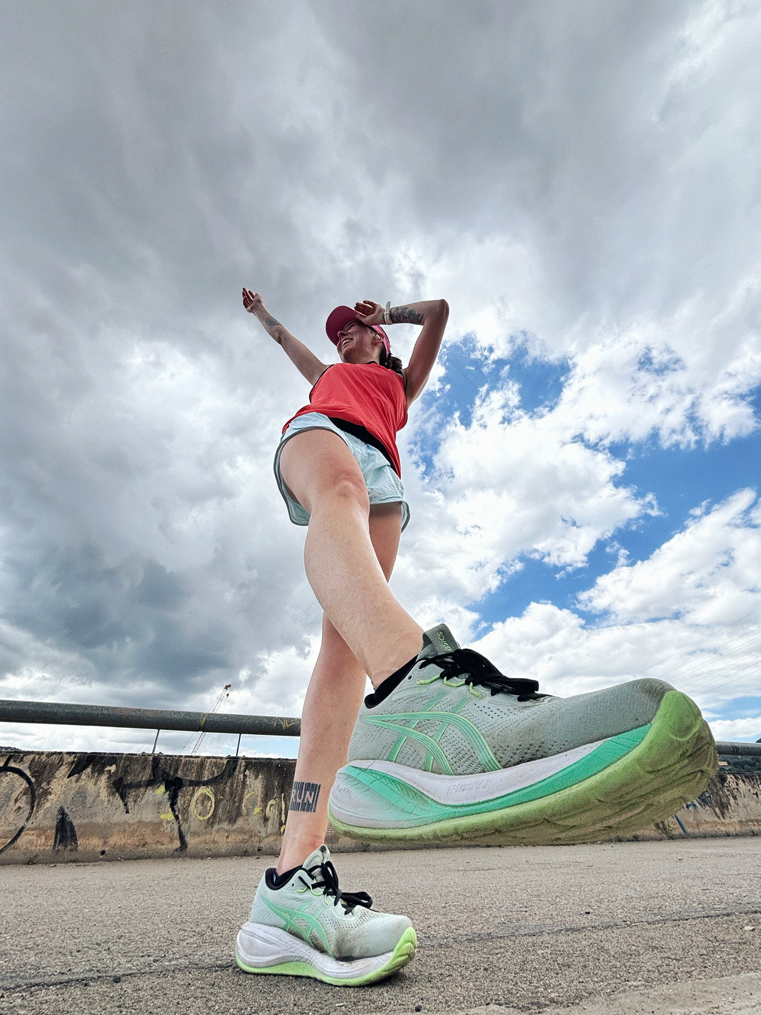 Woman in red top and shorts poses dynamically outdoors wearing ASICS running shoes against cloudy sky.