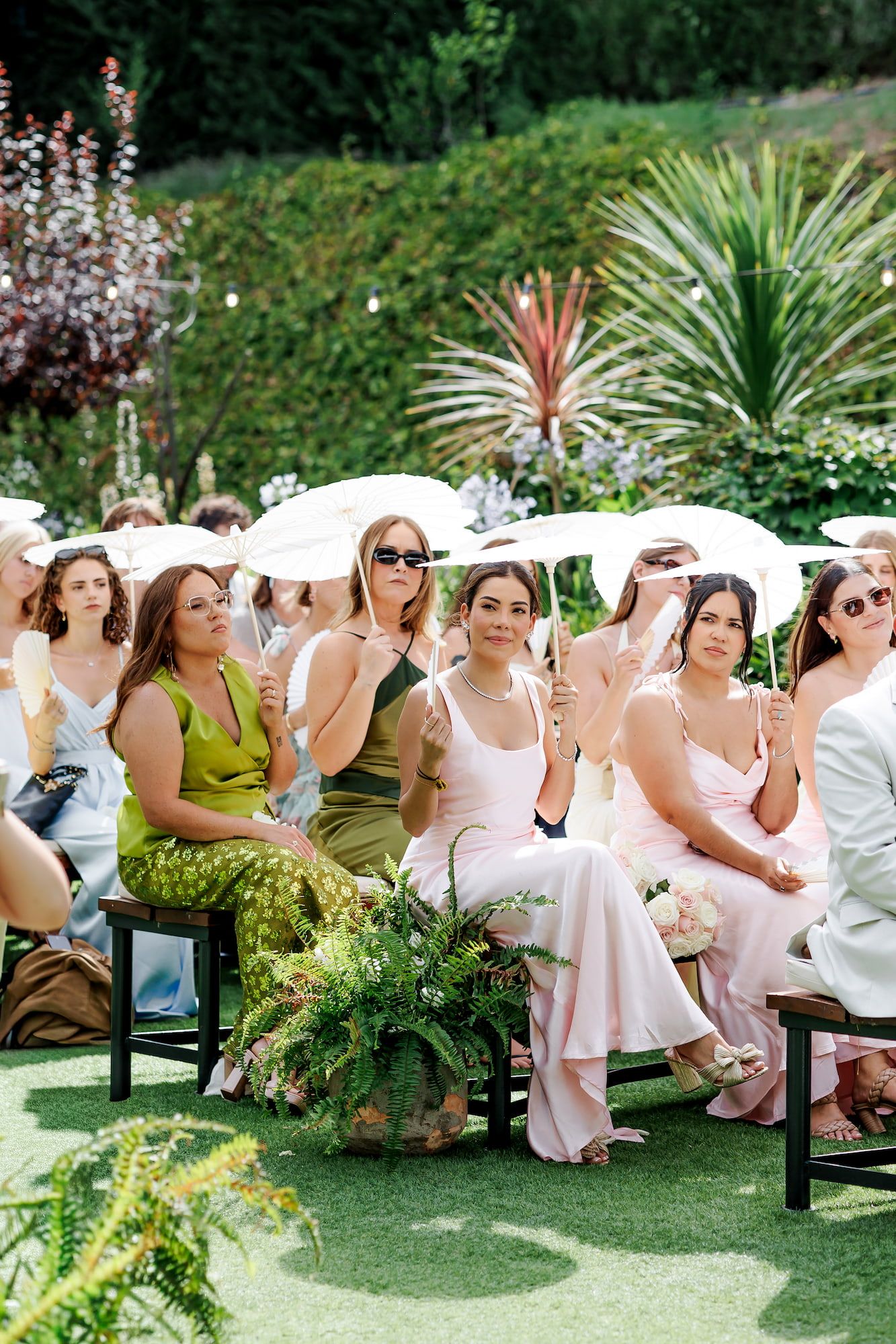 Guests in colorful dresses hold white parasols at an elegant outdoor garden wedding ceremony.