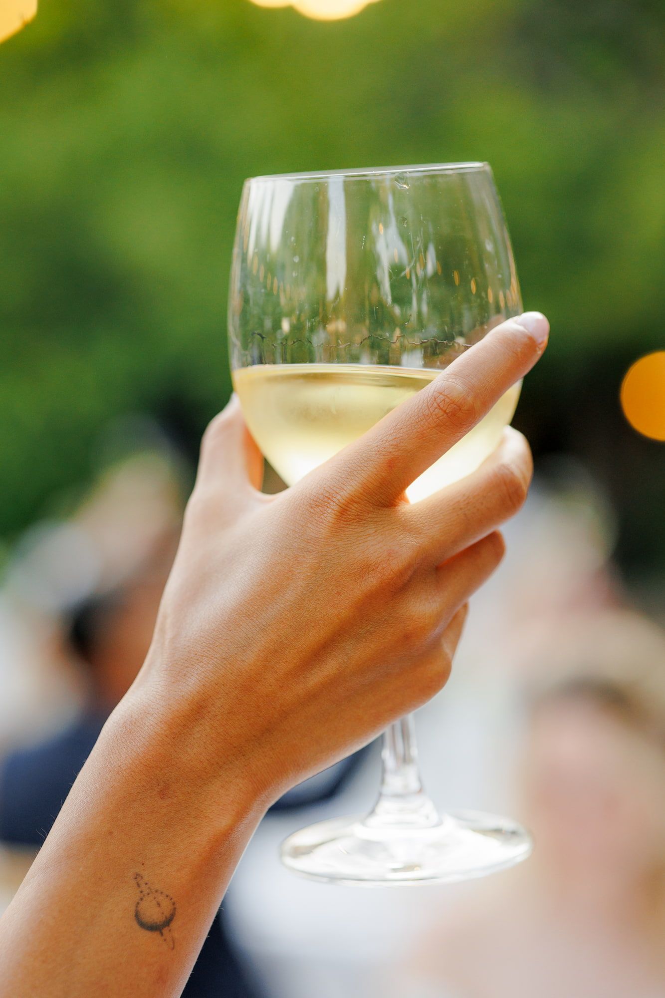 A hand holding a glass of white wine outdoors at a garden party, with a small tattoo visible on the wrist.