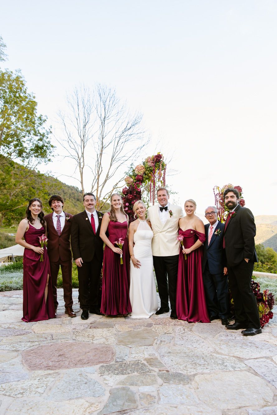 Bride in white gown and groom in ivory jacket pose with bridesmaids in burgundy dresses and groomsmen on a stone patio near a floral arch.