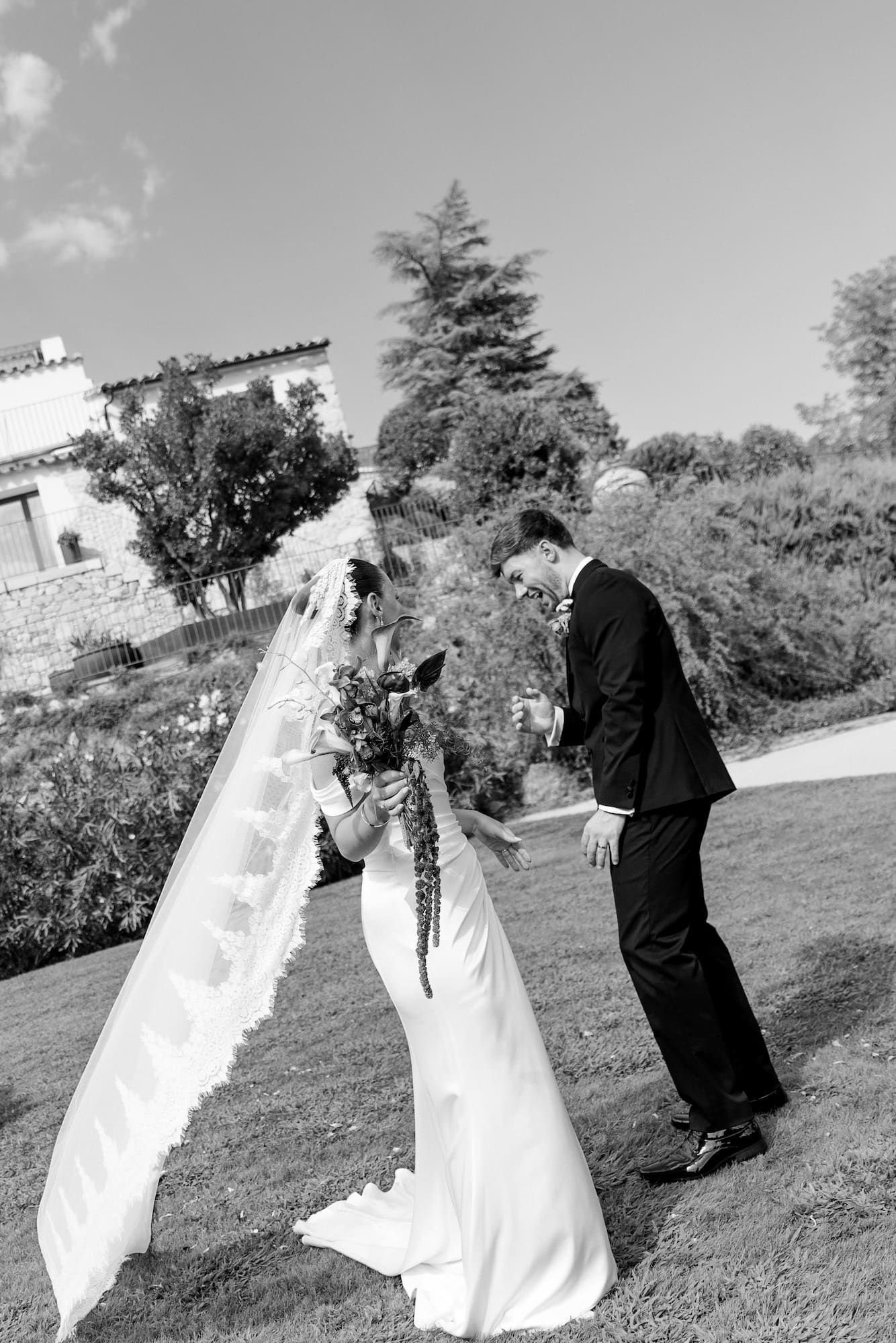 Black and white photo of a bride and groom sharing a tender moment outdoors, with a flowing veil and bouquet.
