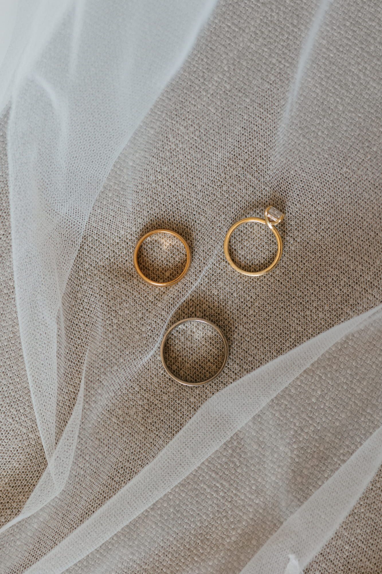 Three gold wedding rings and a diamond engagement ring arranged on a beige linen fabric with a sheer white veil.