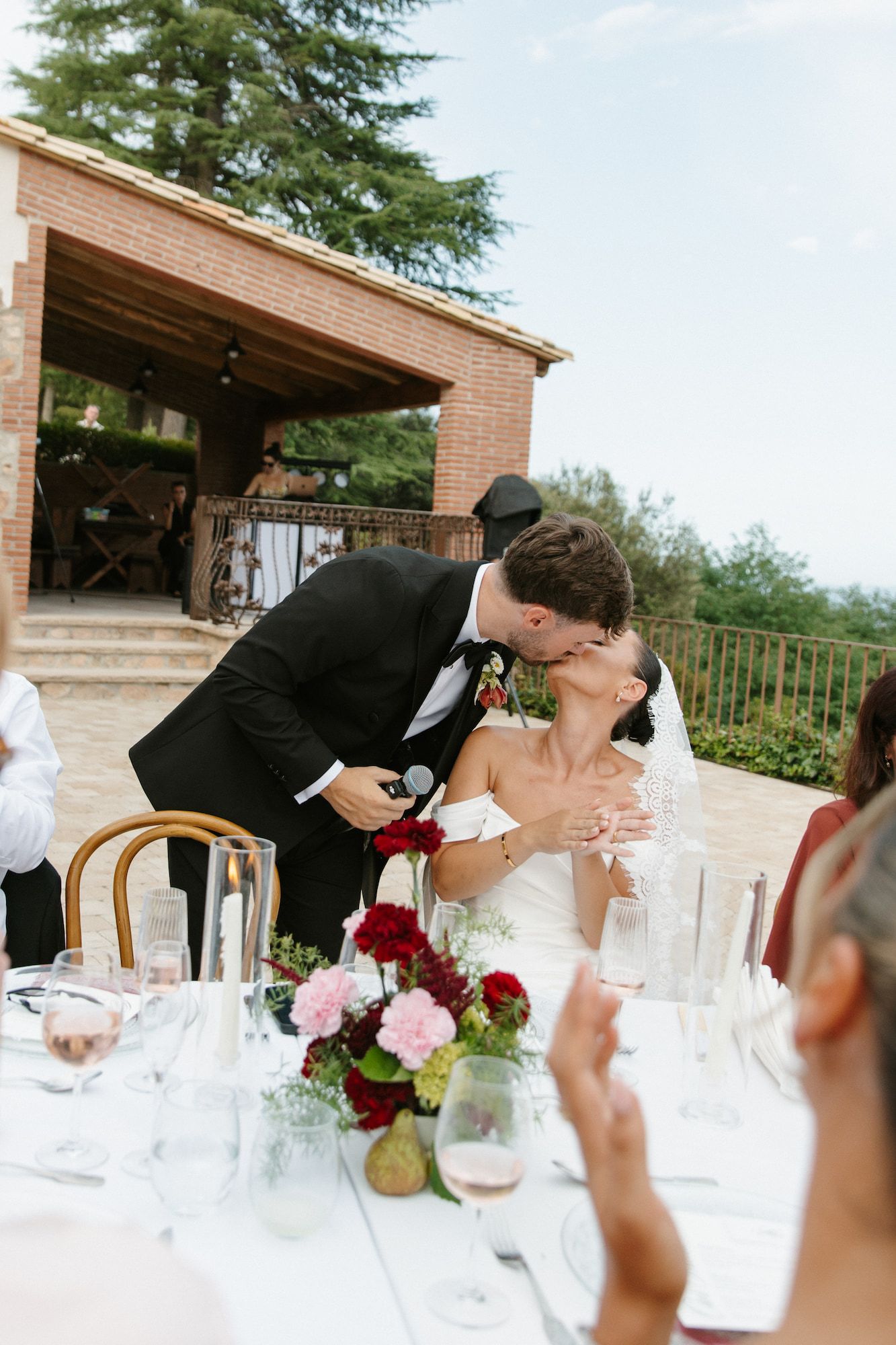 Bride and groom share a kiss at outdoor wedding reception table with floral centerpieces and guests applauding.